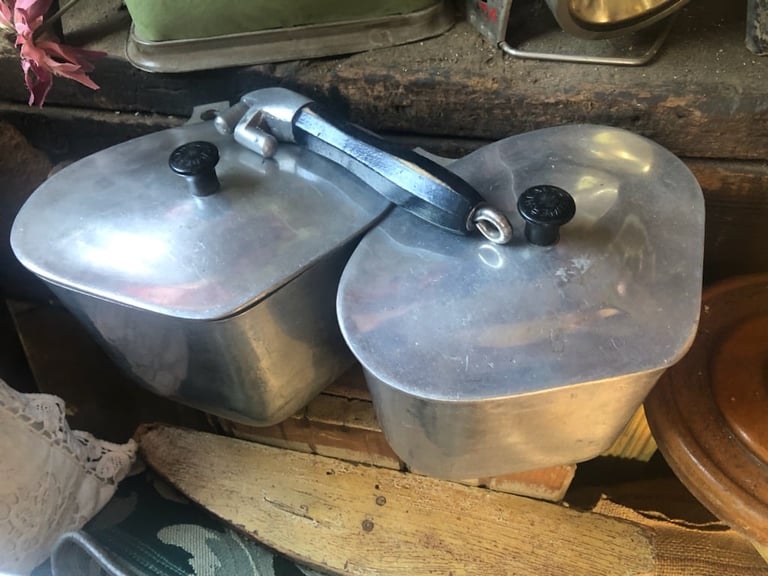 Two stainless steel pressure cookers with silver lids and release valves sitting on a wooden shelf
