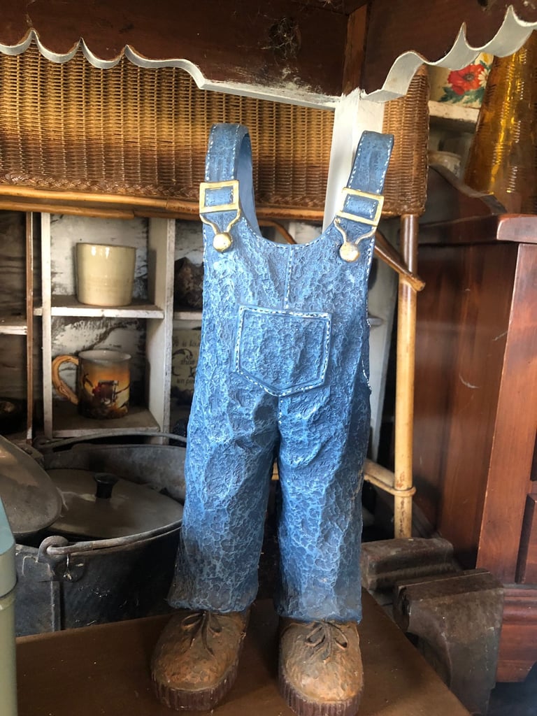 Vintage denim overalls with blue suspenders displayed on a mannequin in a rustic cabin setting with historic furnishings