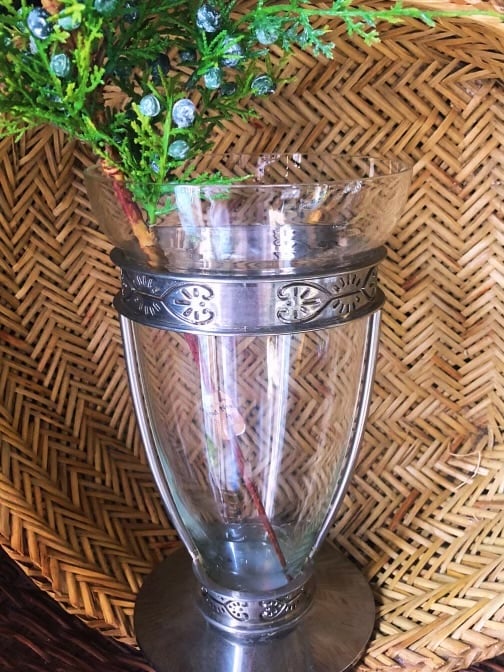 Clear glass vase with decorative silver band holding blue flowering plant on woven basket background