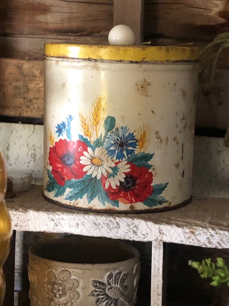 Vintage tin canister with floral design featuring red poppies, daisies, and blue flowers, sitting on a shelf with rustic wooden background