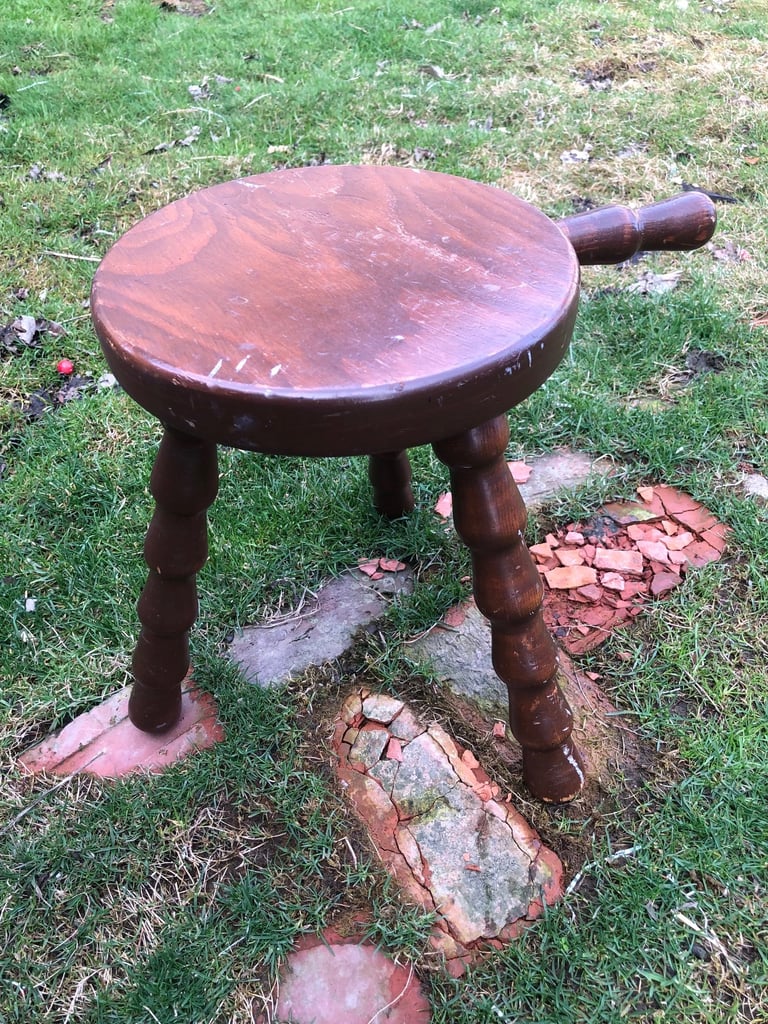 Wooden three-legged stool with round seat and handle on grassy ground with scattered bricks