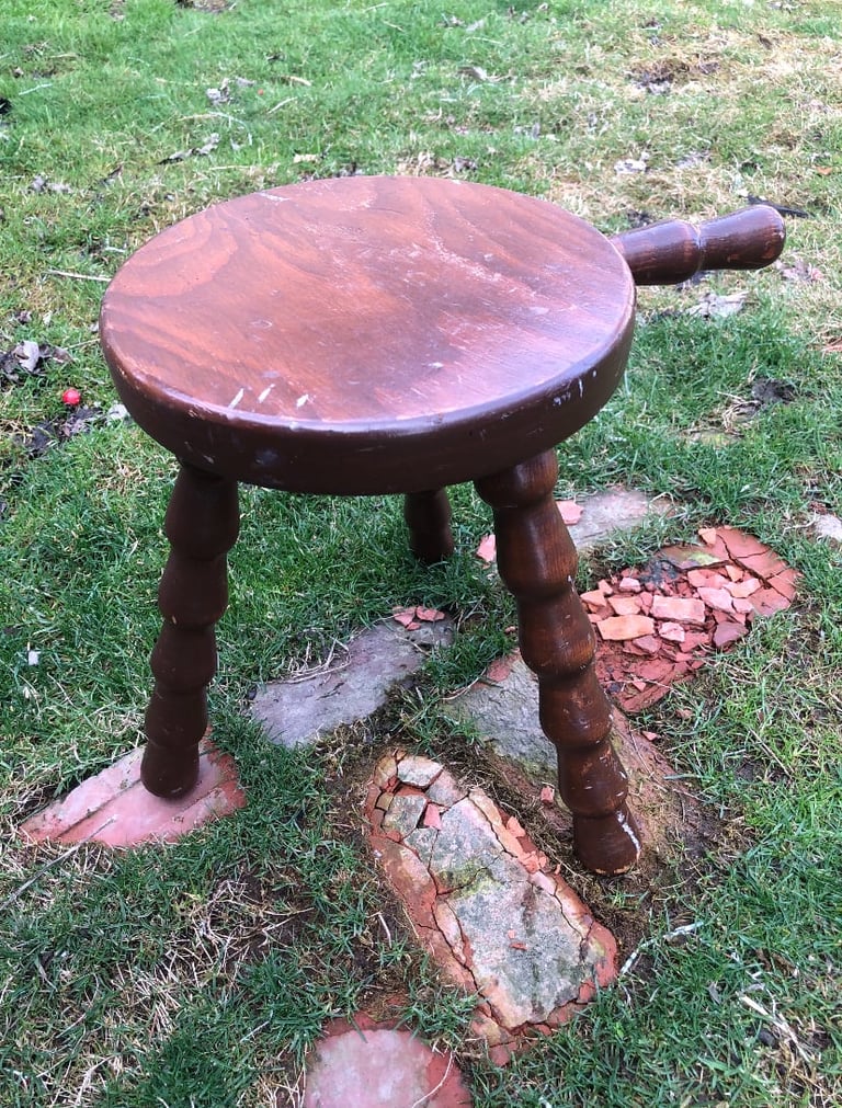 Wooden three-legged stool with round seat and handle, sitting on grass and brick pieces